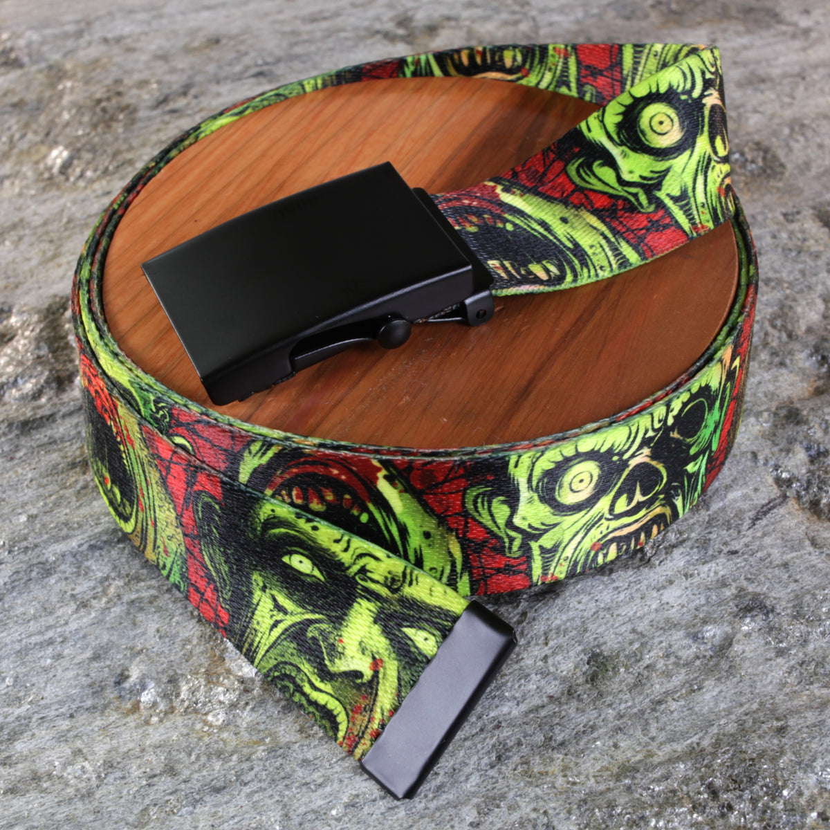 A wide belt with a black buckle, featuring a vibrant green, red, and black horror zombie face print, displayed on a round wooden coaster over a gray stone surface.